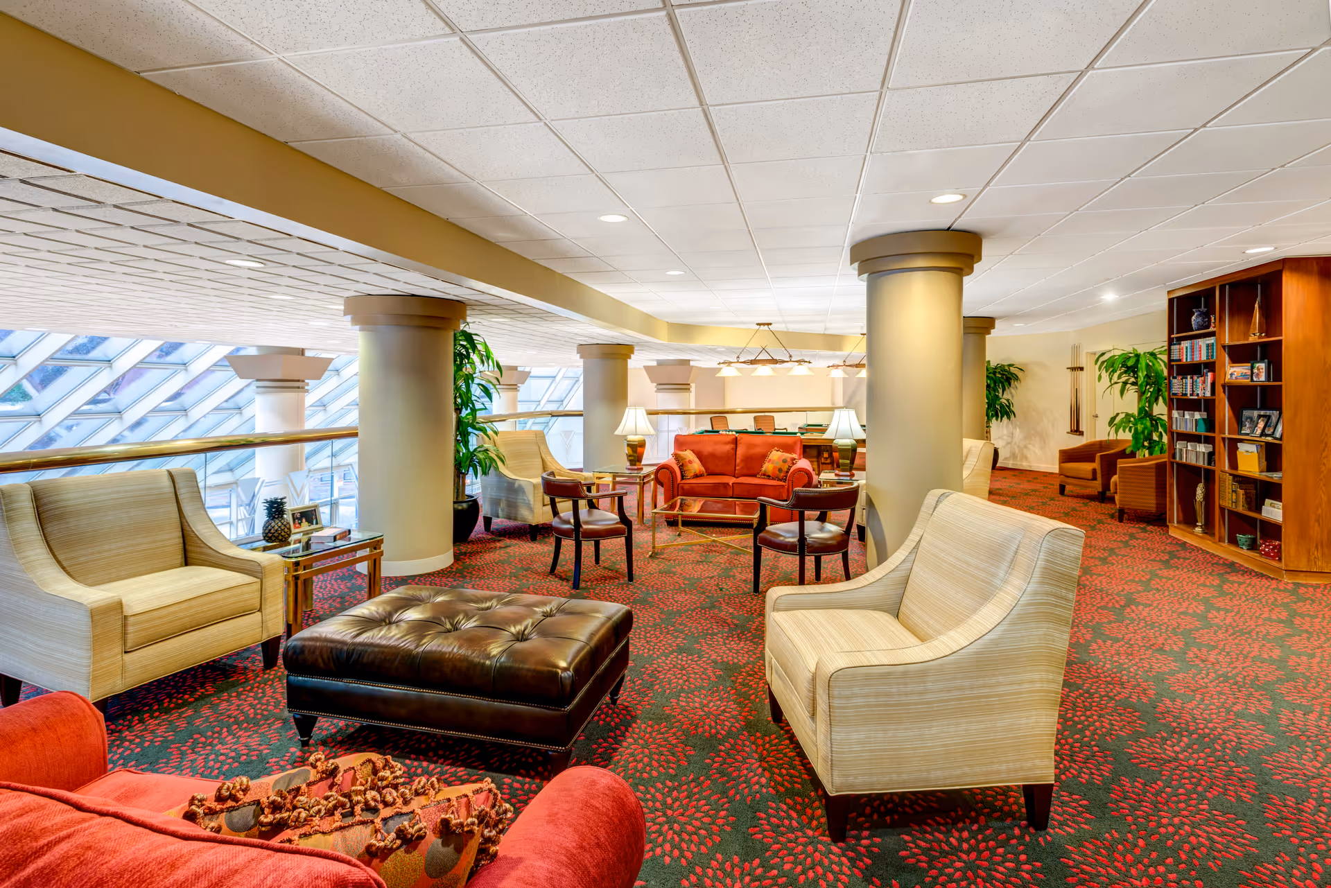 A spacious and well-lit common area with comfortable seating including armchairs, a red sofa, and a large leather ottoman. The room features large windows with a slanted design allowing natural light to fill the space. There are several columns, potted plants, a bookshelf filled with books and decorative items, and table lamps providing additional lighting. The carpet has a red and green floral pattern.