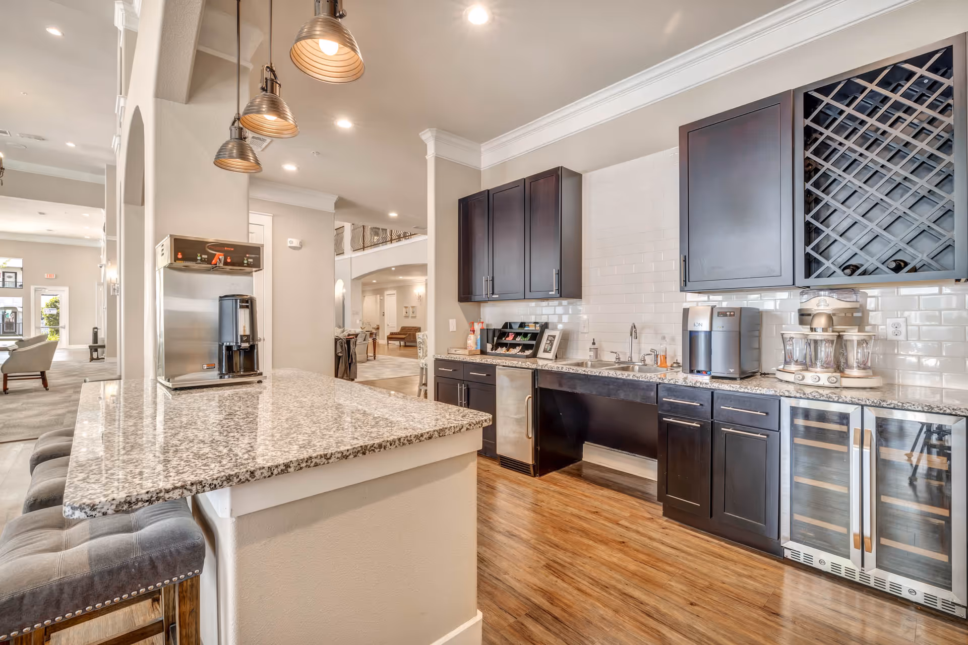 Modern kitchen area with granite countertops, dark wood cabinets, a wine rack, a small sink, and various appliances including a coffee machine and a water dispenser. There are three pendant lights hanging above a breakfast bar with cushioned stools. The kitchen opens into a spacious living area with large windows and comfortable seating.