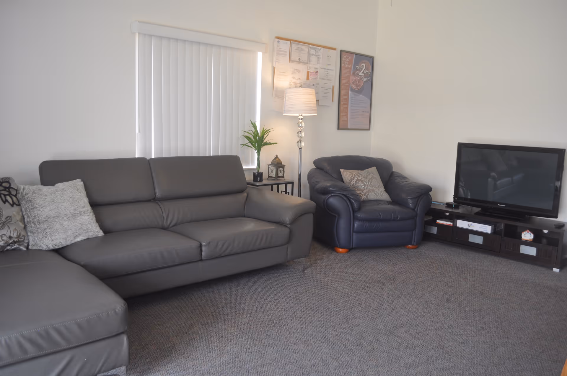 A living room with a gray sectional sofa adorned with decorative pillows, a dark blue armchair with a patterned cushion, a flat-screen TV on a dark wooden TV stand, a side table with a potted plant and a lantern, a floor lamp, and framed documents and posters on the wall.