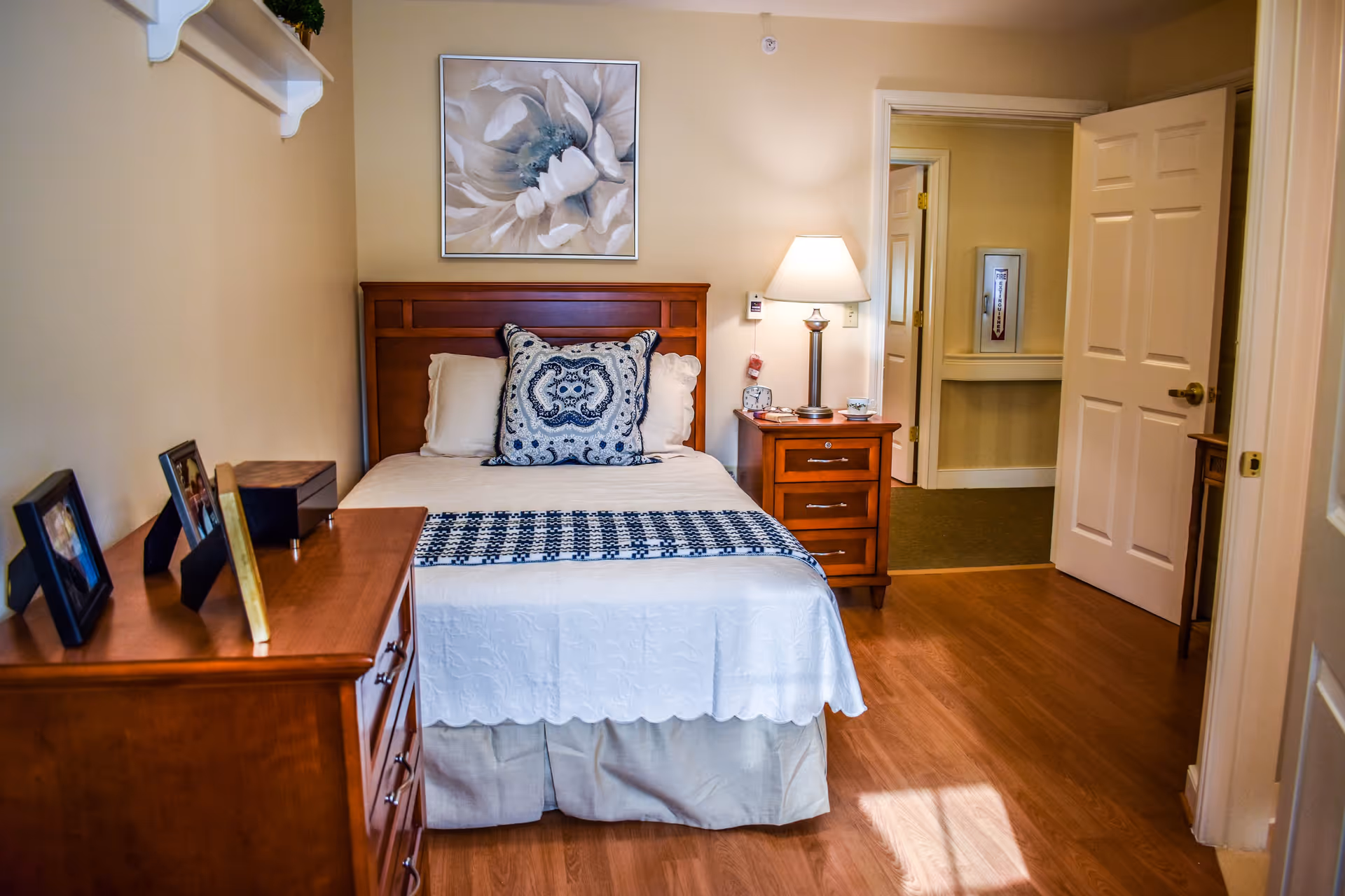 A cozy bedroom with a wooden bed frame, white bedding, and decorative pillows. There is a wooden nightstand with a lamp and alarm clock beside the bed. A wooden dresser with framed photos is on the left side of the room. The walls are painted beige, and a large floral painting hangs above the bed. The door to the room is open, showing a hallway with carpeted flooring.