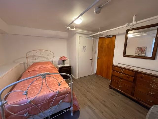 Bedroom with pink-covered metal bed, nightstand with flowers, dresser, and exposed ceiling pipes.