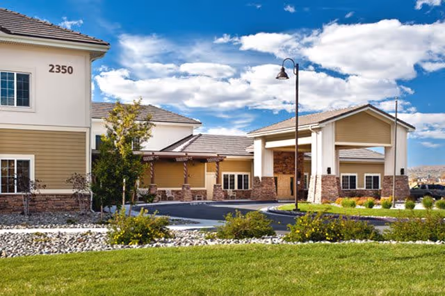 Exterior view of Wingfield Skilled Nursing And Rehabilitation Center showing a modern building with beige and white walls, stone accents, a covered entrance, and a well-maintained lawn under a partly cloudy blue sky.