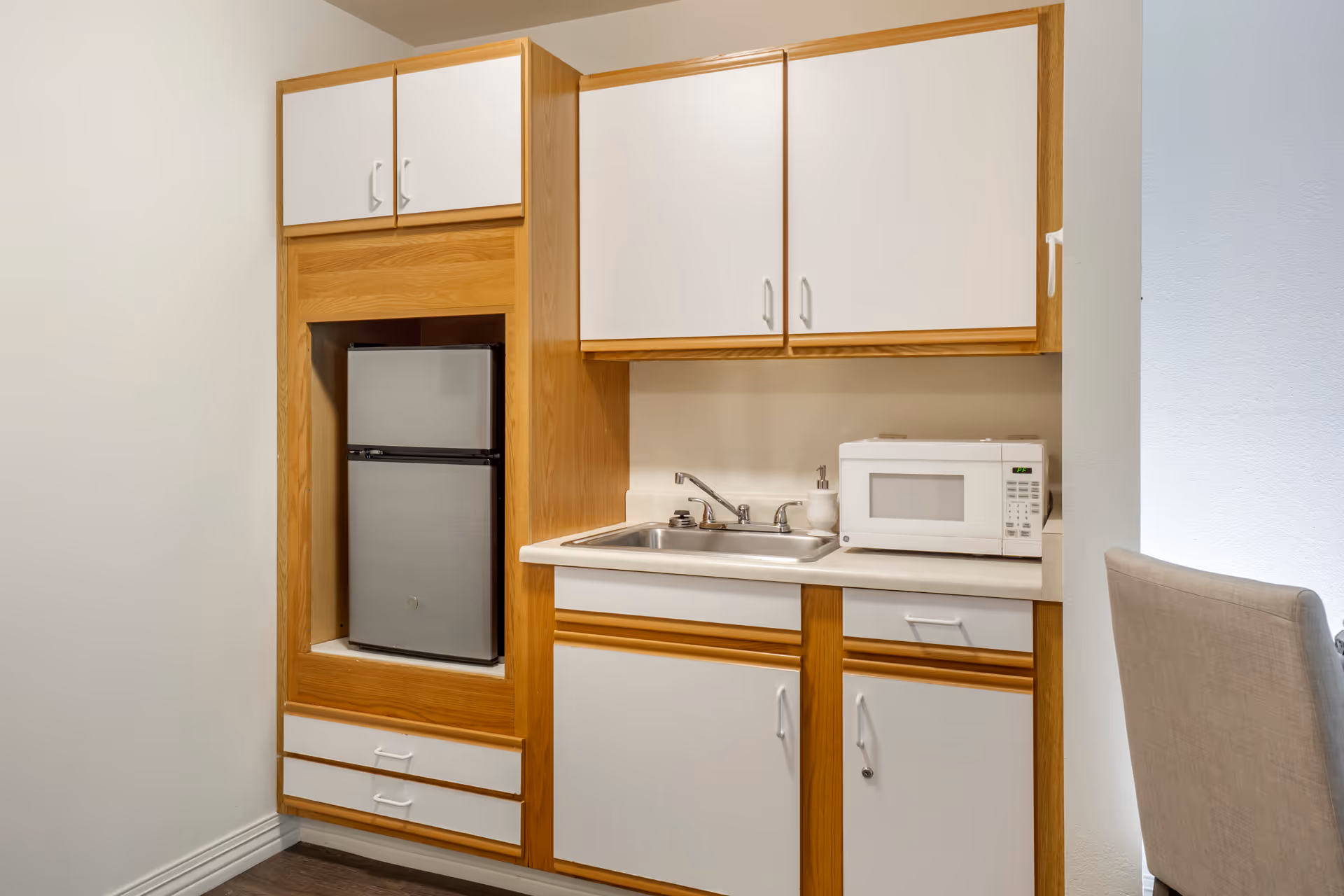 Small kitchenette with wooden and white cabinets, a compact refrigerator, a stainless steel sink, and a white microwave on the countertop. A beige upholstered chair is partially visible on the right side.