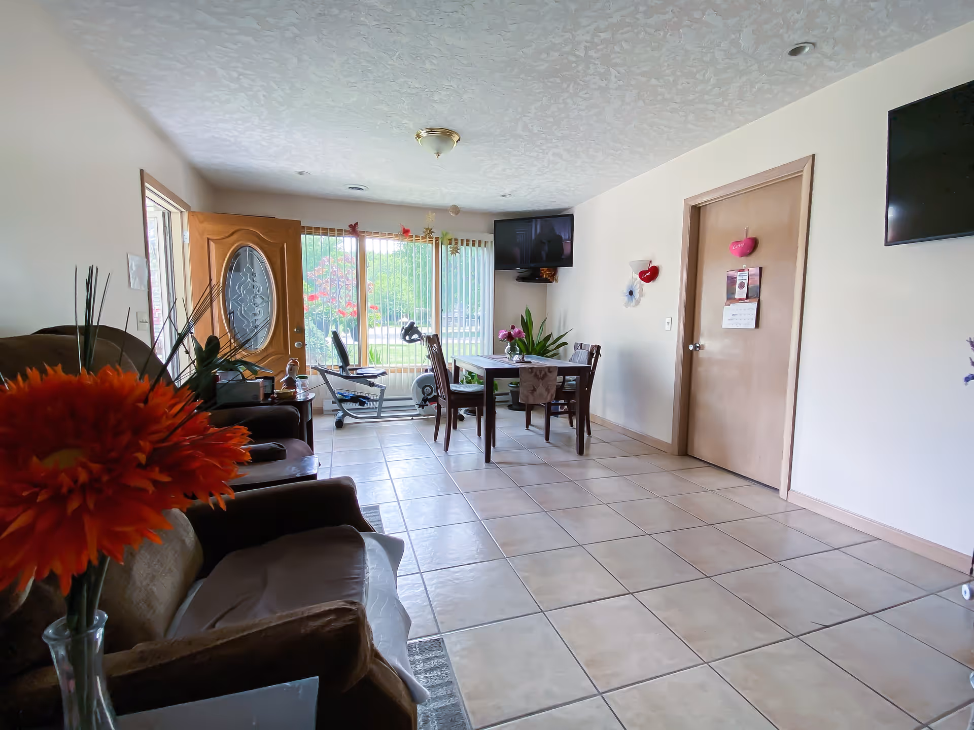 A bright living room area with tiled floors, a wooden front door with glass design, a small dining table with four chairs, two wall-mounted televisions, and a stationary exercise bike near large windows with vertical blinds. There are decorative flowers and plants placed around the room.