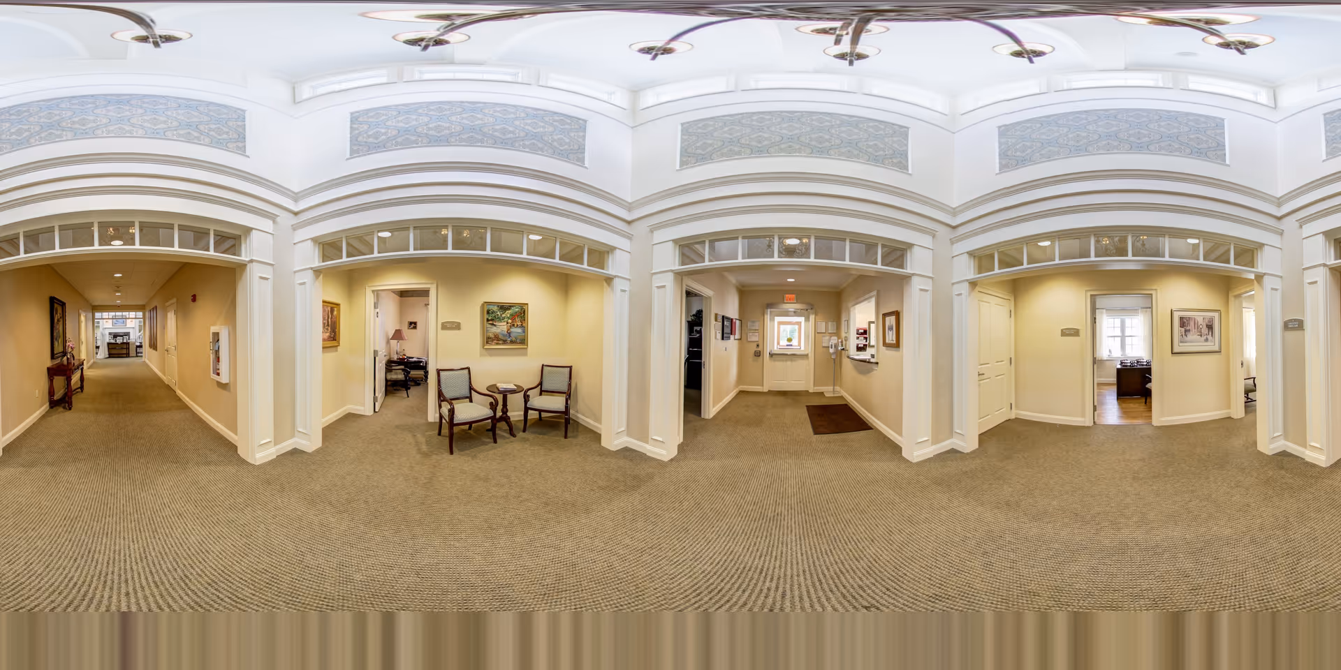 Panoramic view of a carpeted interior hallway/lobby with seating alcoves, decorative archways, and framed artwork.
