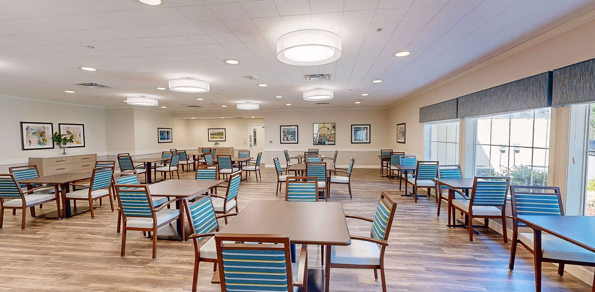 A spacious dining room with multiple square tables and wooden chairs featuring blue and green striped cushions. The room has large windows on one side letting in natural light, light wood flooring, and several framed pictures on the walls. Modern circular ceiling lights illuminate the space.