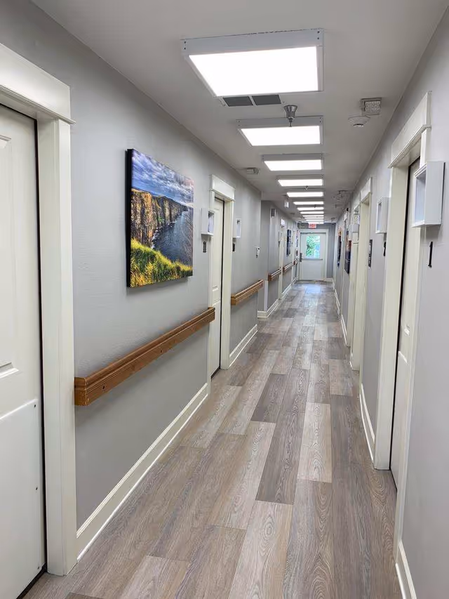 A long, well-lit hallway in a memory care facility with light wood flooring, gray walls, white doors on both sides, wooden handrails, and a landscape painting on the left wall. Ceiling lights illuminate the corridor leading to a door with a window at the far end.