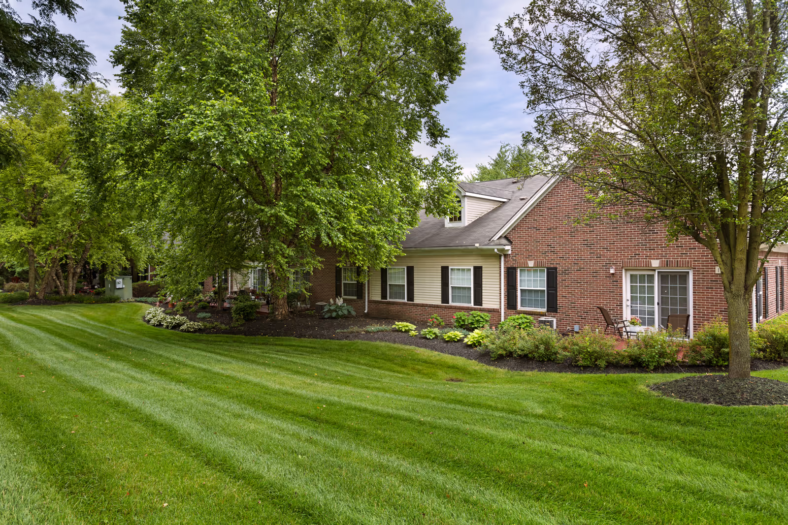 A well-maintained lawn with neatly mowed grass in front of a brick and siding building. The building has several windows with black shutters and a patio area with chairs and a table. There are multiple trees and landscaped garden beds with shrubs and plants surrounding the building.