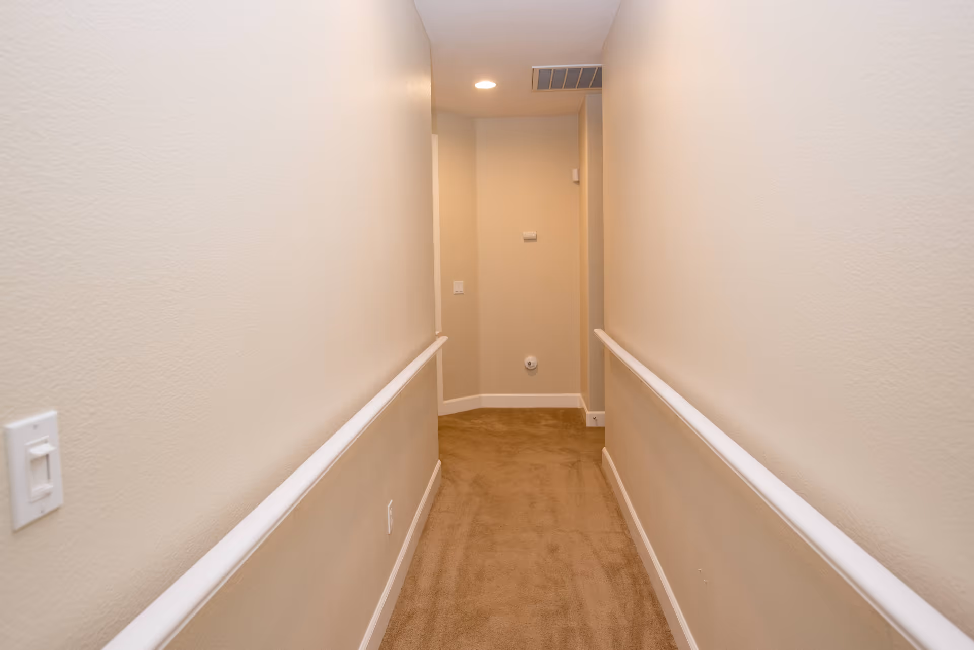 A narrow indoor hallway with beige walls and carpeted floor, featuring white handrails on both sides and a ceiling light at the end.
