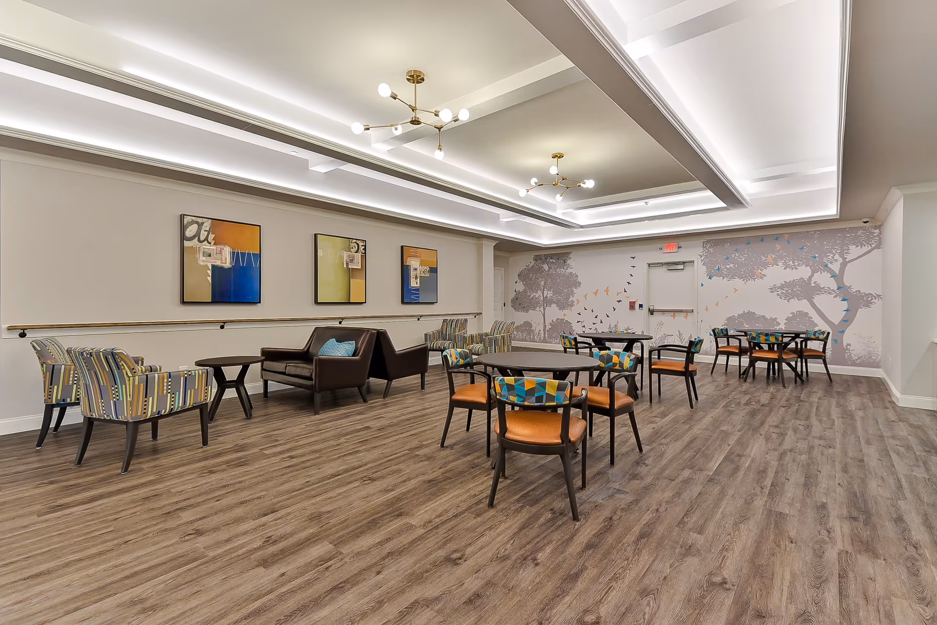 A spacious, well-lit common area with modern ceiling lights, wood flooring, and a variety of seating options including round tables with colorful cushioned chairs and a seating area with armchairs and a leather loveseat. The walls feature abstract artwork and a mural of trees and flying birds.