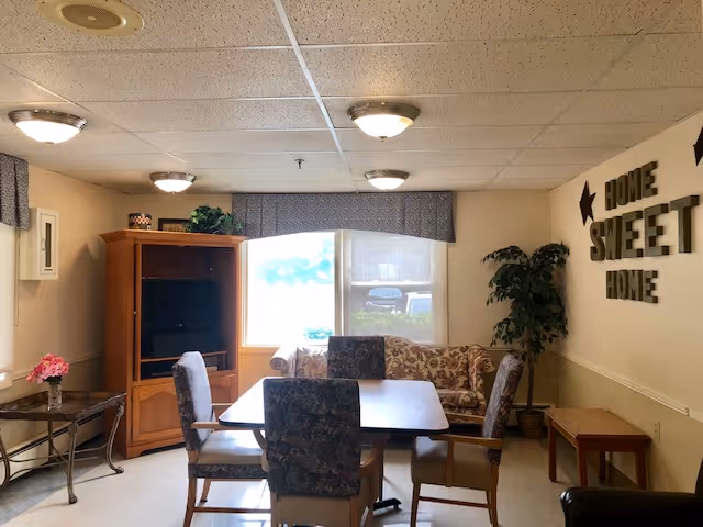 A cozy living room area with a wooden table surrounded by four upholstered chairs. Behind the table is a floral-patterned couch in front of a large window with a valance. To the left, there is a wooden entertainment center with a TV. On the right wall, decorative letters spell out 'HOME SWEET HOME' with star accents. There is a potted plant in the corner and a small wooden bench along the right wall. The ceiling has several round light fixtures.
