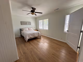 A simple bedroom with a wooden floor, a bed with white bedding against the left wall, two windows letting in natural light, a ceiling fan with light fixture, and a white door on the right side.