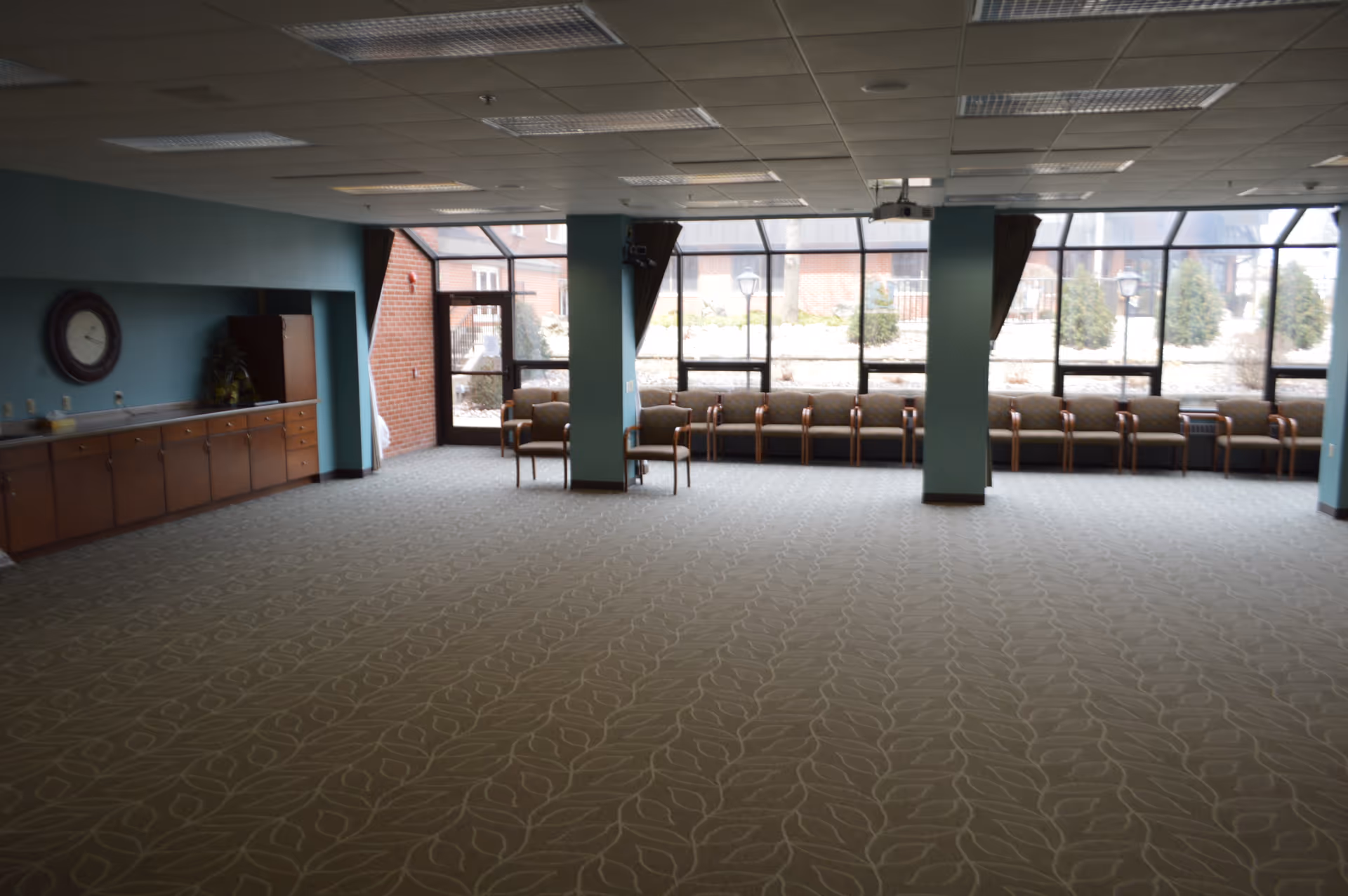 A spacious room with patterned carpet flooring, teal walls, and a row of chairs lined up against large windows that let in natural light. There is a wooden cabinet with a clock on the wall above it on the left side, and two teal pillars near the windows. The room appears to be a common area or lounge in an assisted living facility.