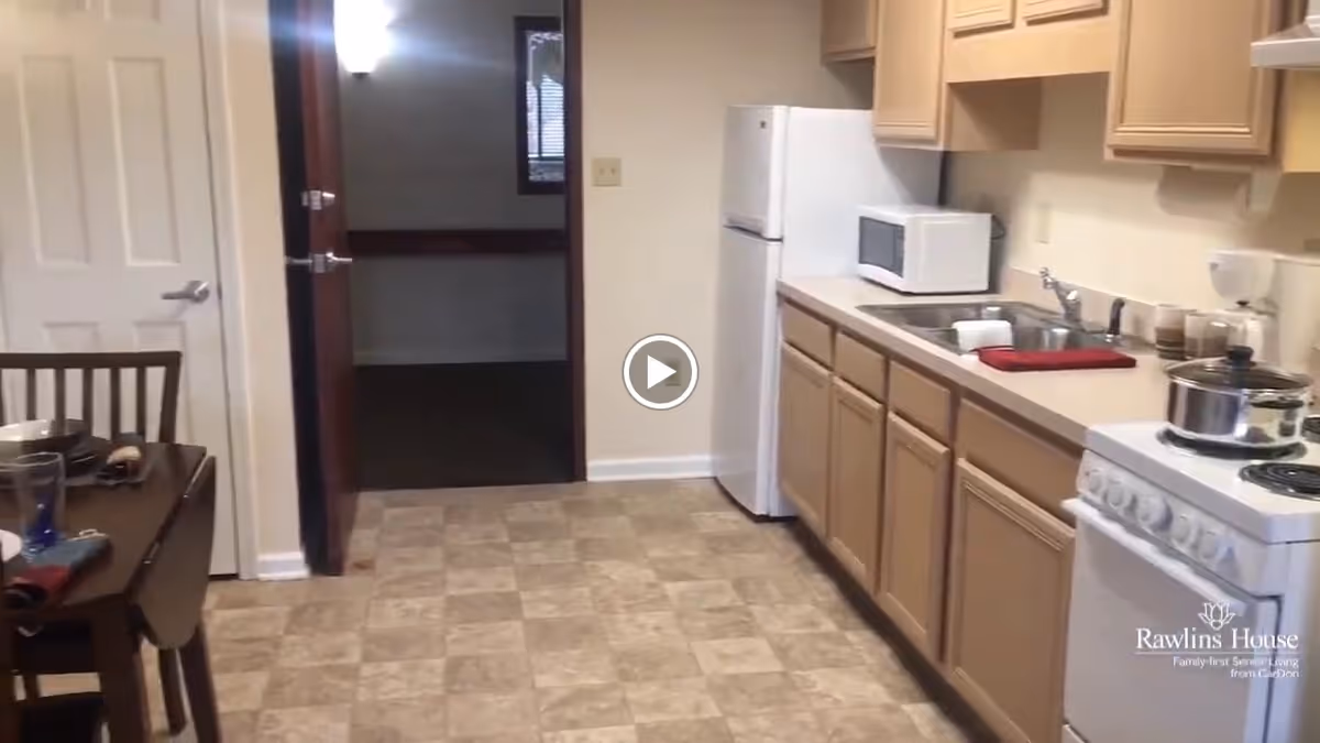 Interior view of a kitchen in a senior living facility featuring beige cabinets, a white refrigerator, microwave, stove with a pot on it, and a sink with a red towel. To the left, there is a dining table with chairs and place settings. The floor is covered with beige patterned tiles and there is an open doorway leading to another room.
