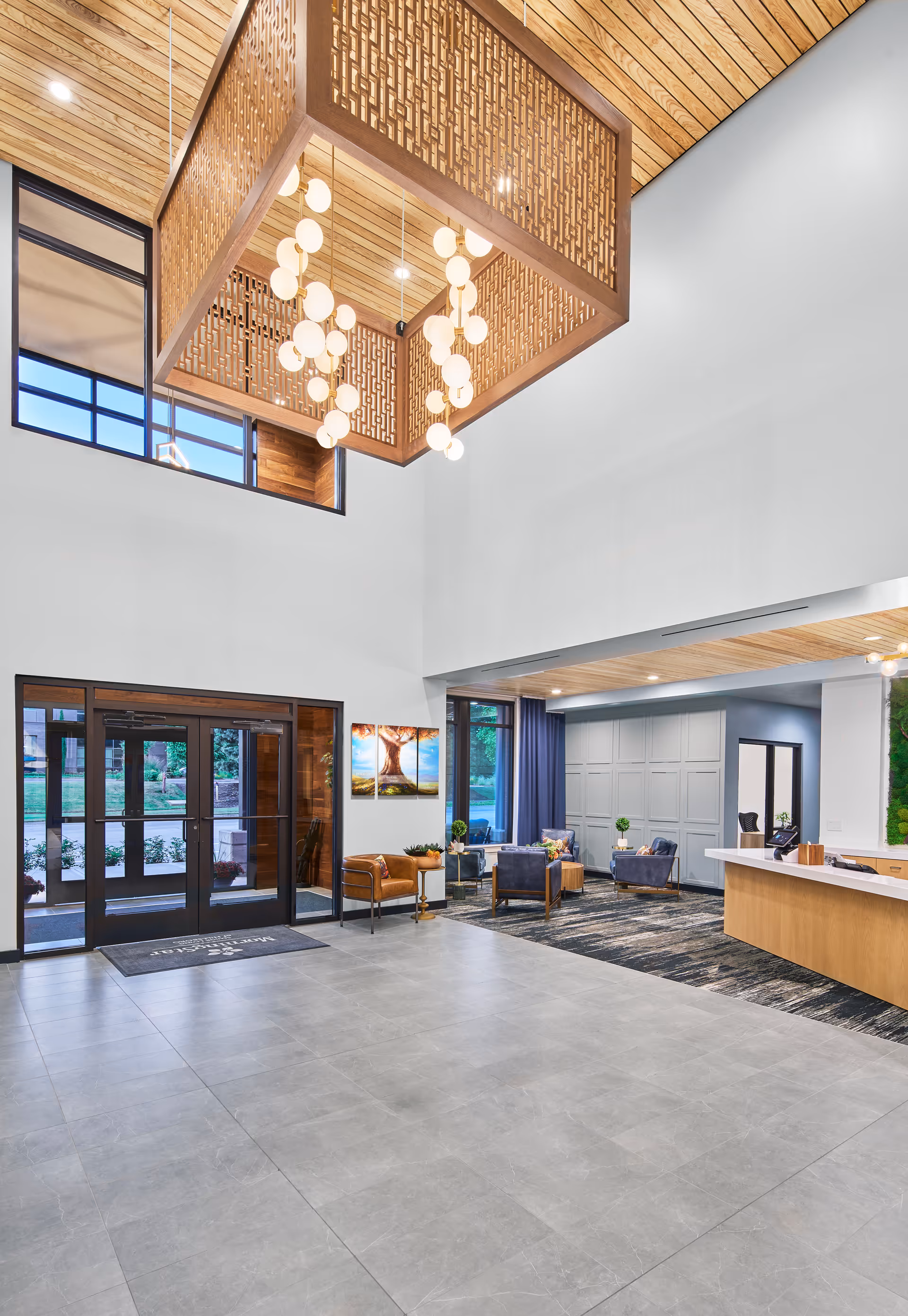 Spacious and modern senior living facility lobby with large gray tiled floor, wooden ceiling, and a decorative wooden light fixture hanging from the ceiling. The lobby features glass double doors, comfortable seating areas with chairs and small tables, a reception desk, and large windows allowing natural light to enter.