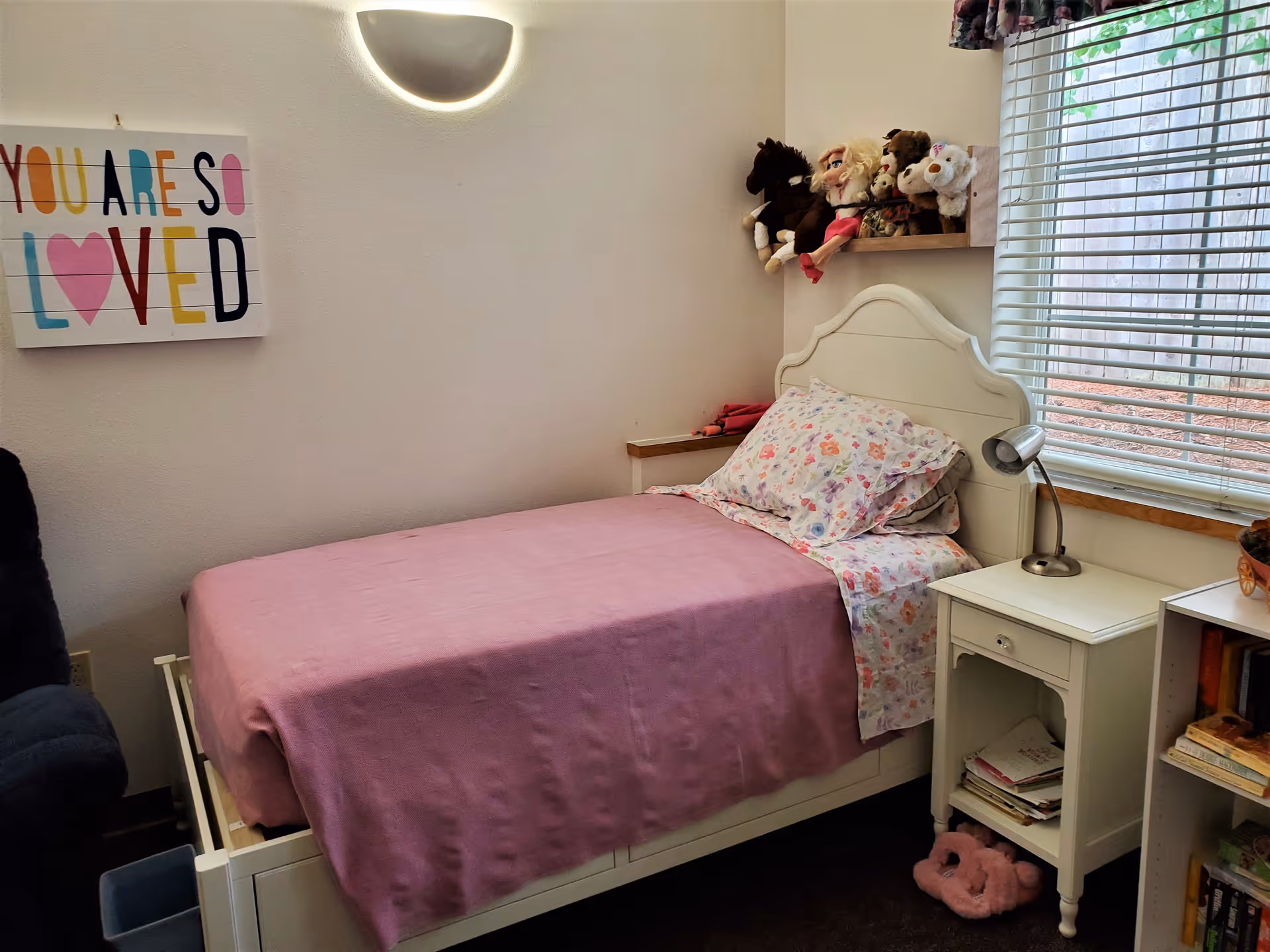 A small bedroom with a single bed covered in a pink blanket and floral pillowcases. Above the bed is a shelf holding several stuffed animals. To the right of the bed is a white nightstand with a small lamp and some papers underneath. A window with blinds is on the right wall, and a colorful wall art with the words 'YOU ARE SO LOVED' is hung on the left wall. A wall-mounted light fixture is above the bed.