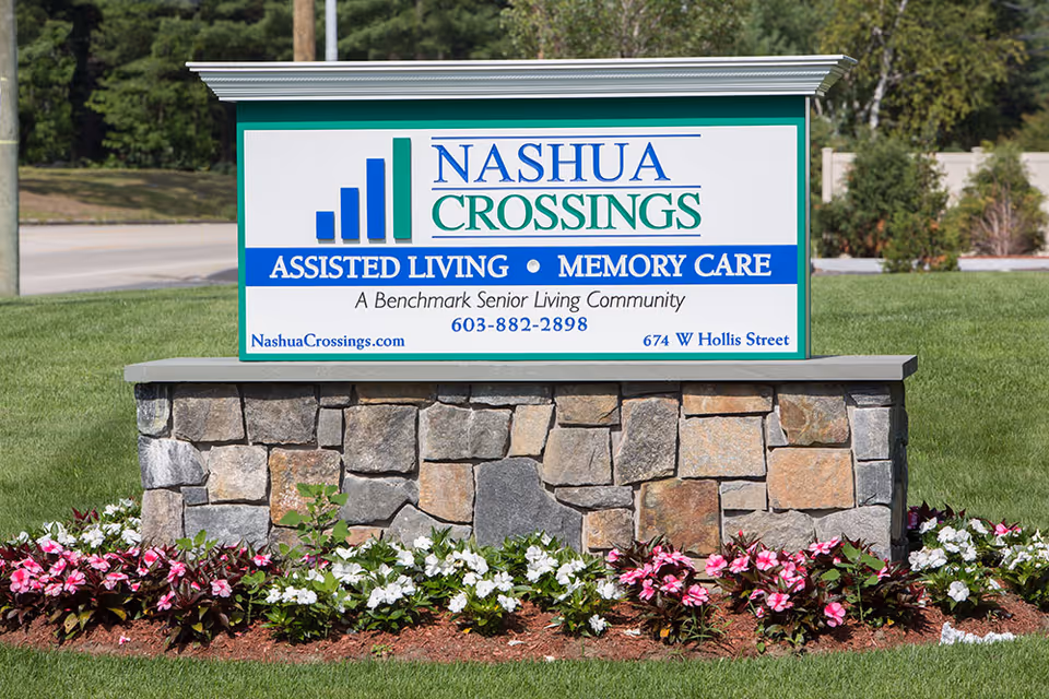Outdoor stone sign for Nashua Crossings Assisted Living and Memory Care, a Benchmark Senior Living Community, surrounded by green grass and pink and white flowers.