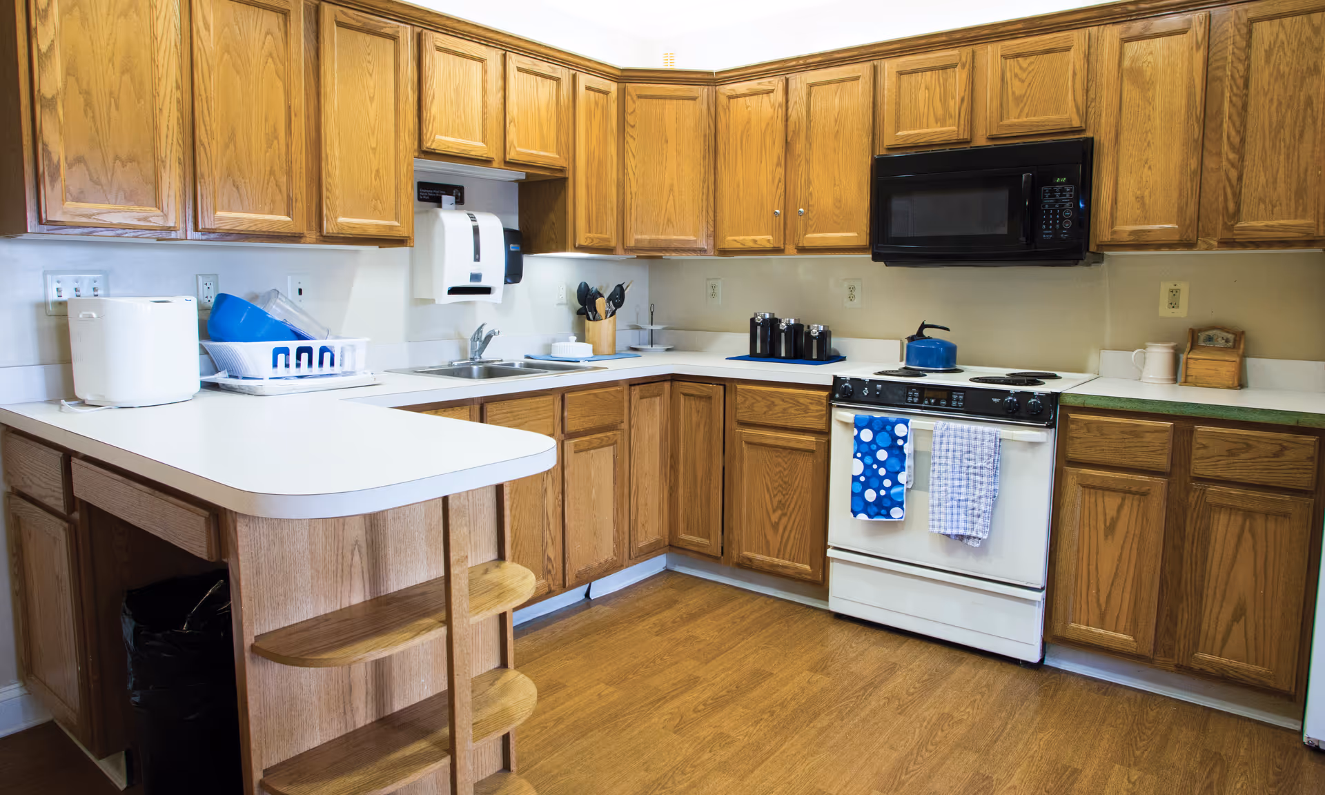 A kitchen with wooden cabinets, white countertops, and a wooden floor. The kitchen features a white stove with a blue kettle on top, a black microwave mounted above the stove, a double sink, a dish rack with dishes, and various kitchen utensils and containers on the counters. There are two towels hanging on the stove handle, one blue with white circles and one white with a blue checkered pattern.
