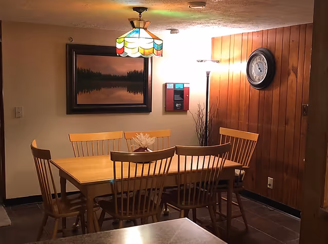 Wooden dining table with six chairs beneath a stained-glass pendant light in a wood-paneled dining area with a wall clock and framed landscape.
