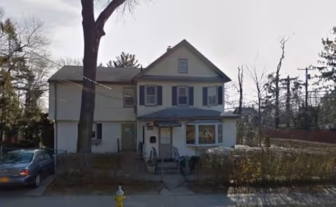 Two-story beige house labeled Manor Haven Adult Home with shrubs and a parked car