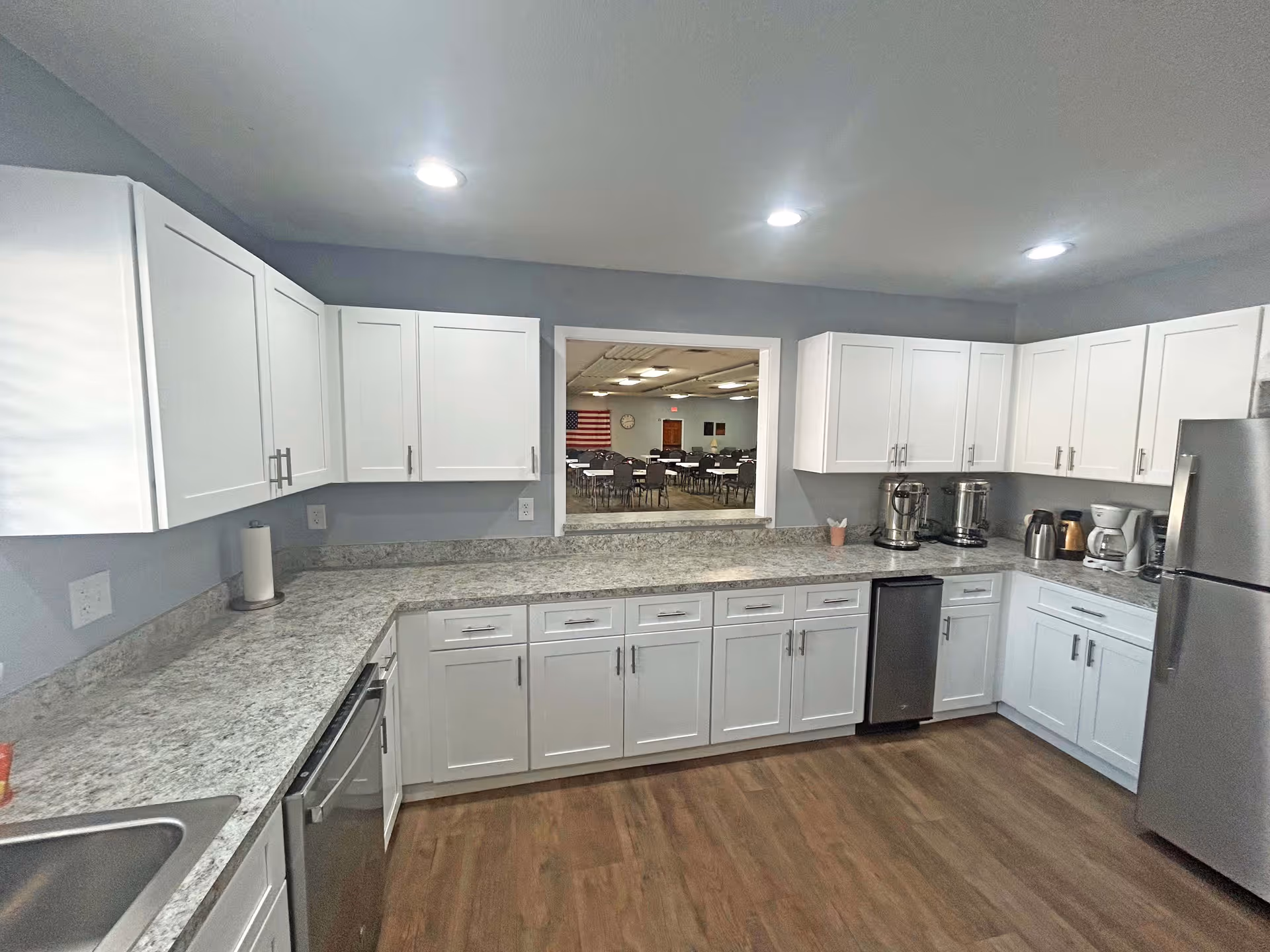 A modern kitchen with white cabinets, gray granite countertops, stainless steel appliances including a refrigerator and dishwasher, and a window opening into a dining area with tables and chairs. The kitchen has recessed lighting and wood flooring.