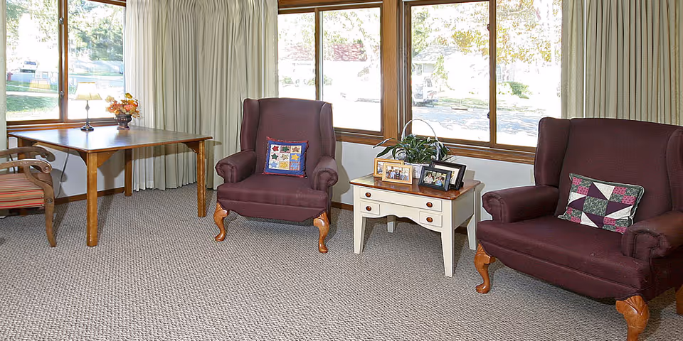 Sunlit sitting area with two maroon wingback chairs, a small side table with framed photos and a wooden table by the window.