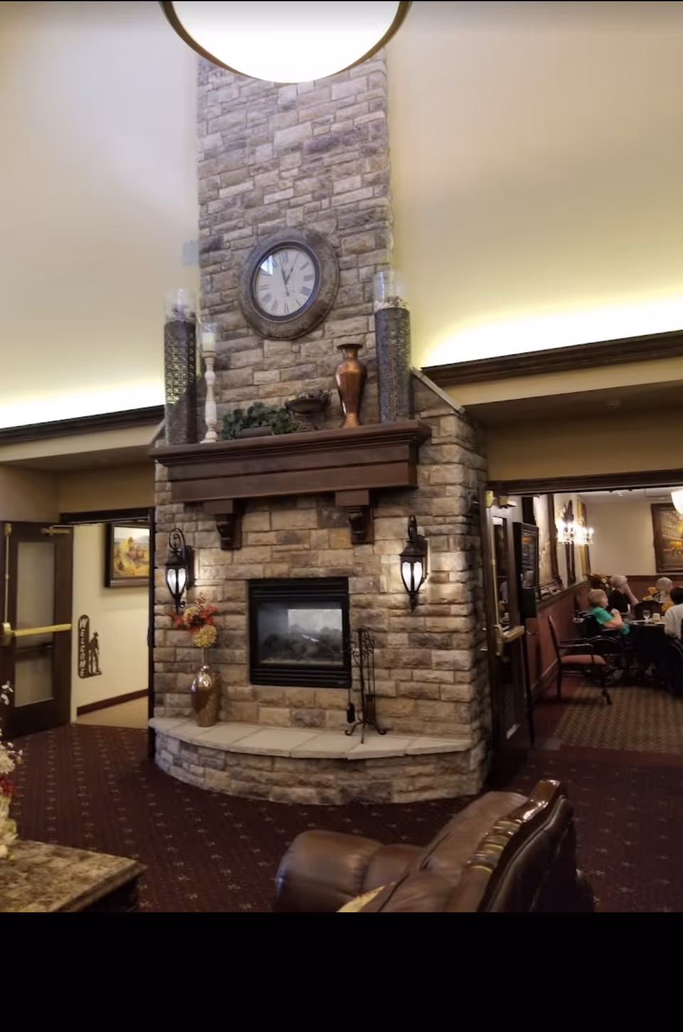 Stone fireplace with a mantel, clock and decorative vases in a communal seating area with a dining room visible to the right.