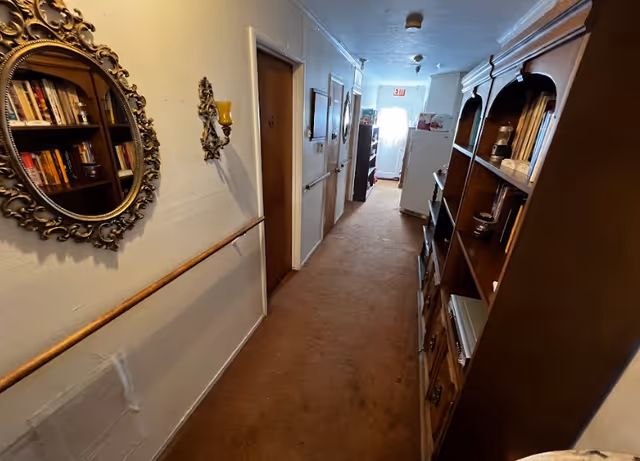 Narrow hallway inside a senior living facility with wall decor, doors, and wooden shelving.