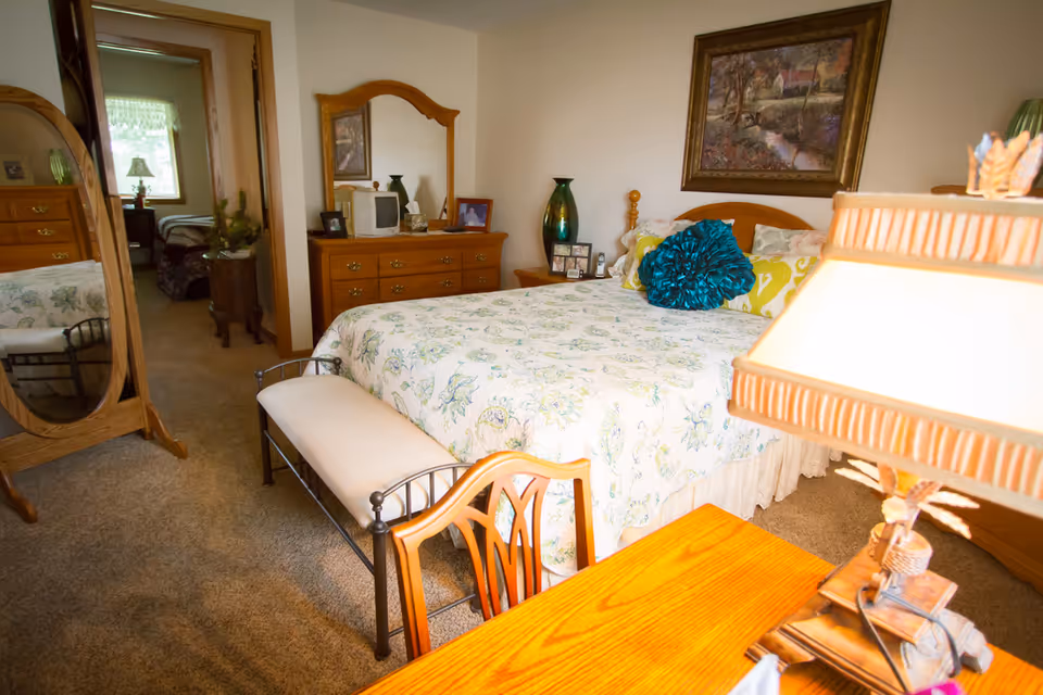 A cozy bedroom with a large bed covered in a floral bedspread and decorative pillows. There is a wooden dresser with a mirror, a small TV, and framed photos on top. A wooden chair and a lamp on a wooden table are in the foreground. A full-length mirror stands to the left, and an open doorway leads to another room with a window and additional furniture.
