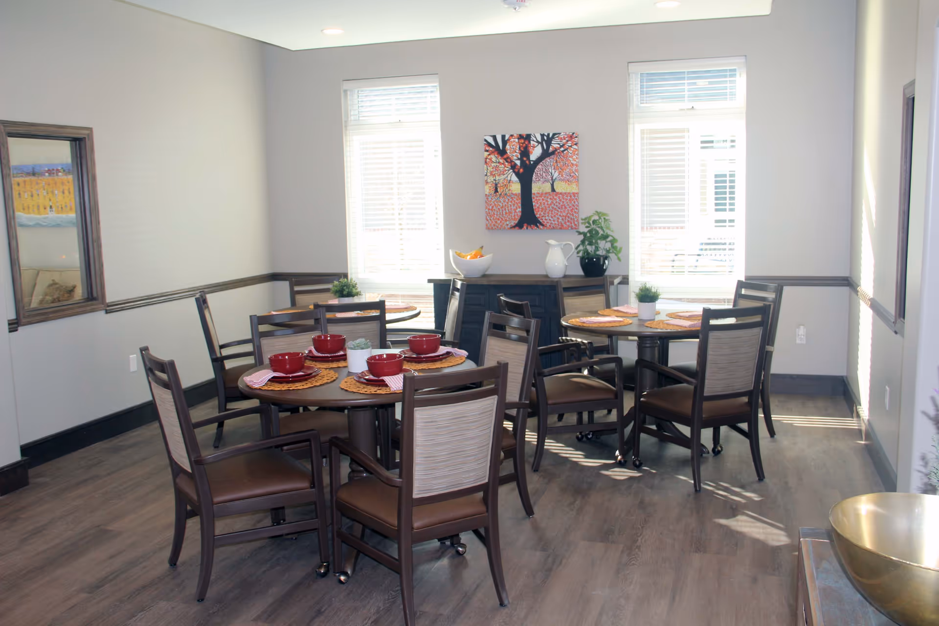 A bright dining room with two round tables, each set with four chairs. The tables have orange placemats, red bowls, and folded napkins. There are two large windows with white blinds letting in natural light. A sideboard against the wall holds a white pitcher, a bowl of fruit, and a potted plant. A colorful painting of a tree with red and orange leaves hangs above the sideboard. The room has light gray walls with dark wood trim and a wood floor.