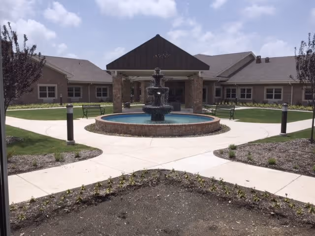 Outdoor courtyard area of a senior living facility featuring a central multi-tiered water fountain surrounded by a circular brick base. The courtyard has paved walkways, benches, small trees, and landscaped garden beds with a building in the background under a partly cloudy sky.