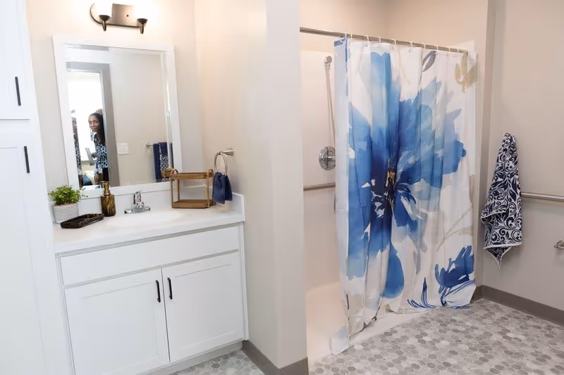A clean and modern bathroom featuring a white vanity with a sink, a mirror above it, and a small plant and basket on the counter. To the right is a shower area with a curtain decorated with large blue flowers. The floor has a hexagonal tile pattern, and there are grab bars installed near the shower and on the wall.