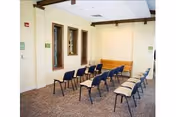 A small meeting or presentation room with two rows of chairs facing a wooden bench against a beige wall. The room has carpeted flooring, two windows on the left wall, and a white ceiling with exposed wooden beams.