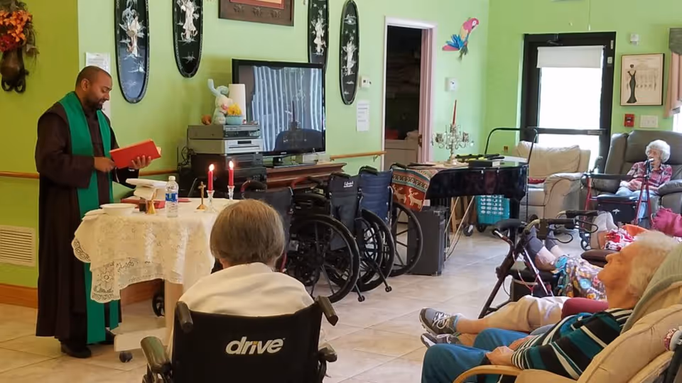 A man dressed in religious attire stands behind a table covered with a white lace cloth, holding a red book and conducting a service in a room with elderly residents seated in wheelchairs and recliners. The room has light green walls, a TV, and various decorations including candles on the table and wall hangings.