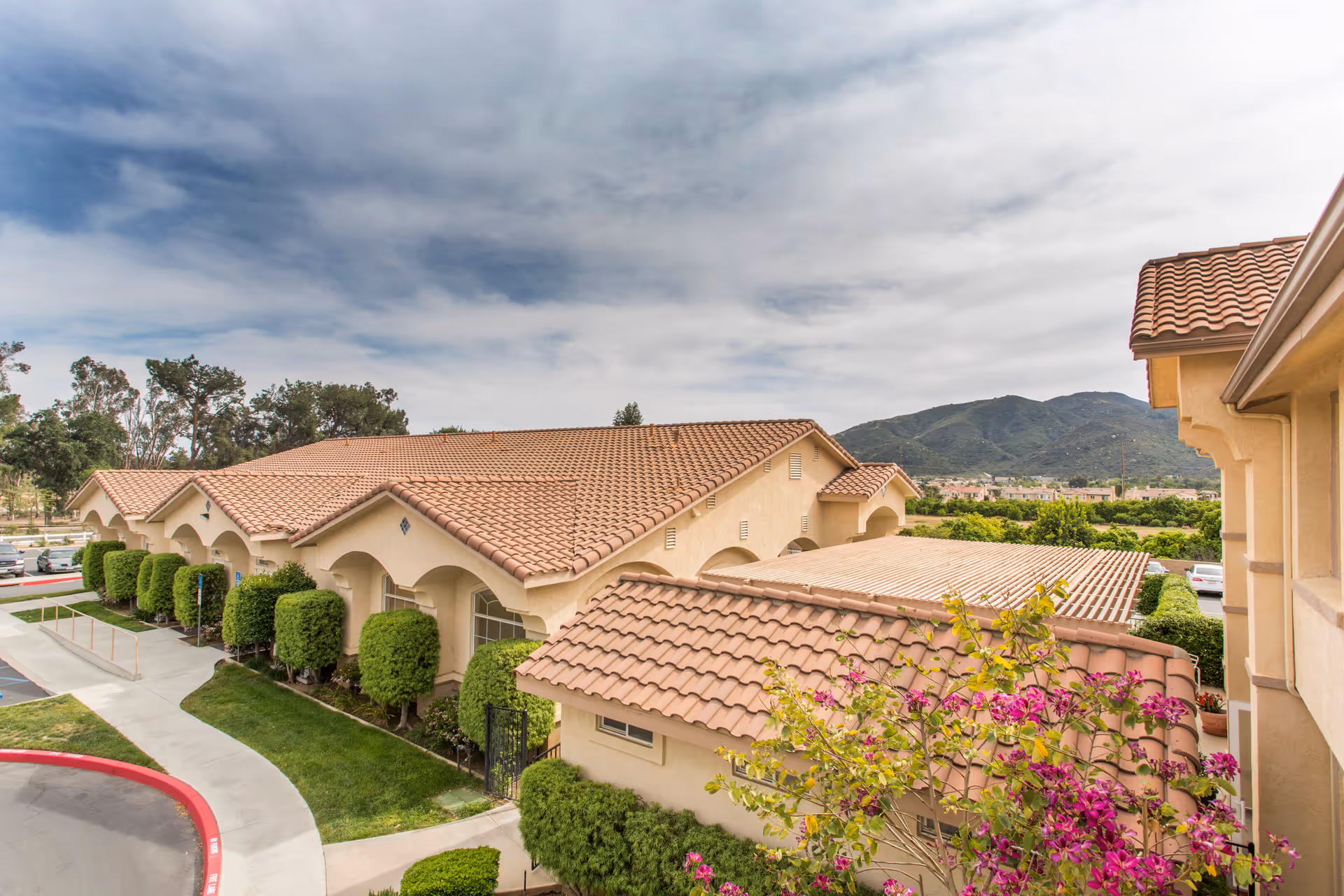 Exterior view of a senior living facility with beige stucco walls and red tile roofs, surrounded by neatly trimmed bushes and trees, with mountains visible in the background under a partly cloudy sky.