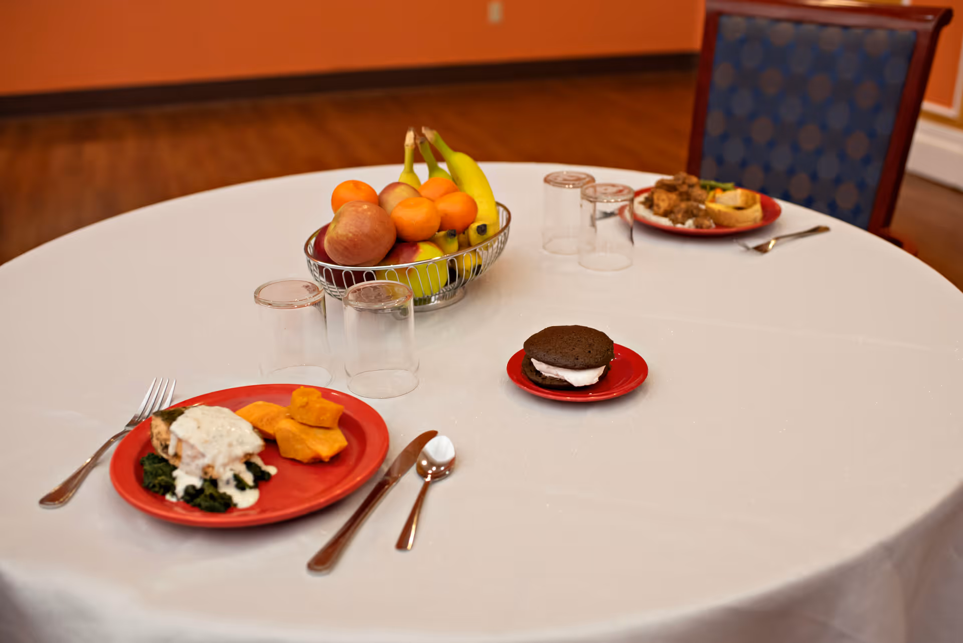 A round table covered with a white tablecloth set with two red plates of food, including a dessert sandwich on a small red plate, and a metal fruit basket filled with bananas, apples, and oranges. There are also three empty drinking glasses and silverware on the table. The background shows an orange wall and a blue cushioned chair.