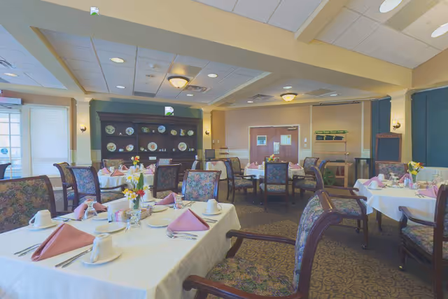 A dining room in an assisted living facility with multiple tables set for a meal. Each table is covered with a white tablecloth, pink folded napkins, cups, glasses, and silverware. Floral centerpieces decorate the tables. The room has floral upholstered chairs, a carpeted floor, and a wooden cabinet displaying decorative plates against the far wall. Soft lighting comes from ceiling fixtures and wall sconces.