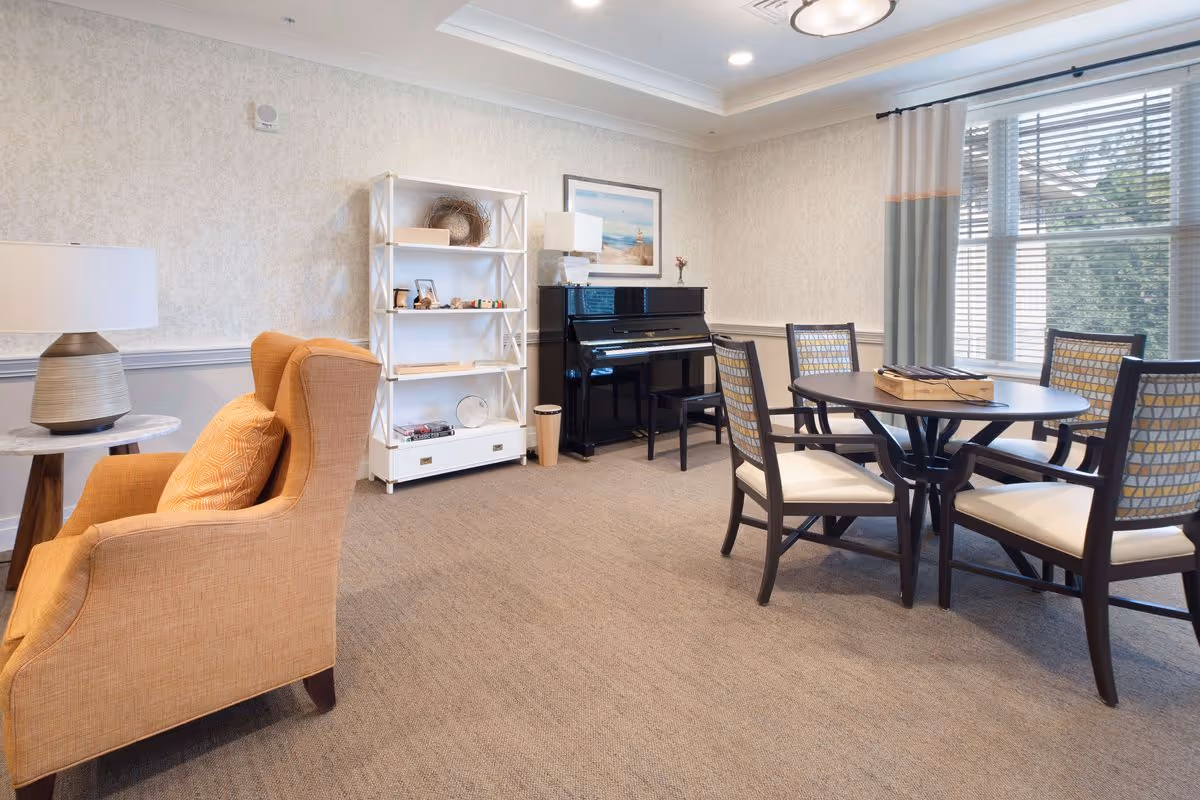 A bright community living room with an orange armchair, round dining table and chairs, a piano, bookshelf, and large windows with blinds.
