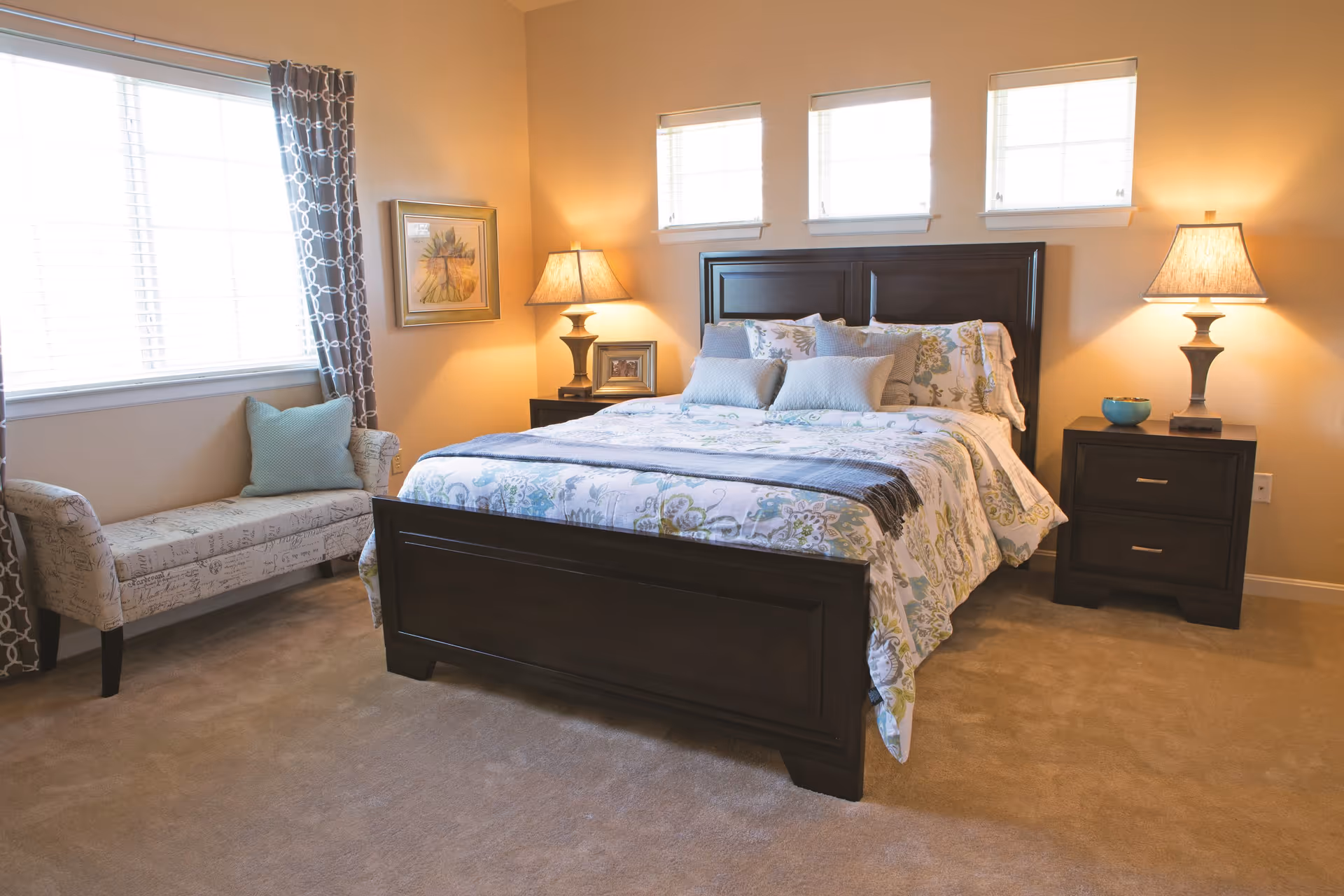 A cozy bedroom with a large dark wooden bed frame, floral bedding, and multiple pillows. There are two matching dark wooden nightstands on either side of the bed, each with a lamp. Three small windows are above the bed, and a larger window with patterned curtains is on the left side of the room. A cushioned bench with a decorative pillow is placed near the window. The walls are painted a warm beige color, and the floor is carpeted.