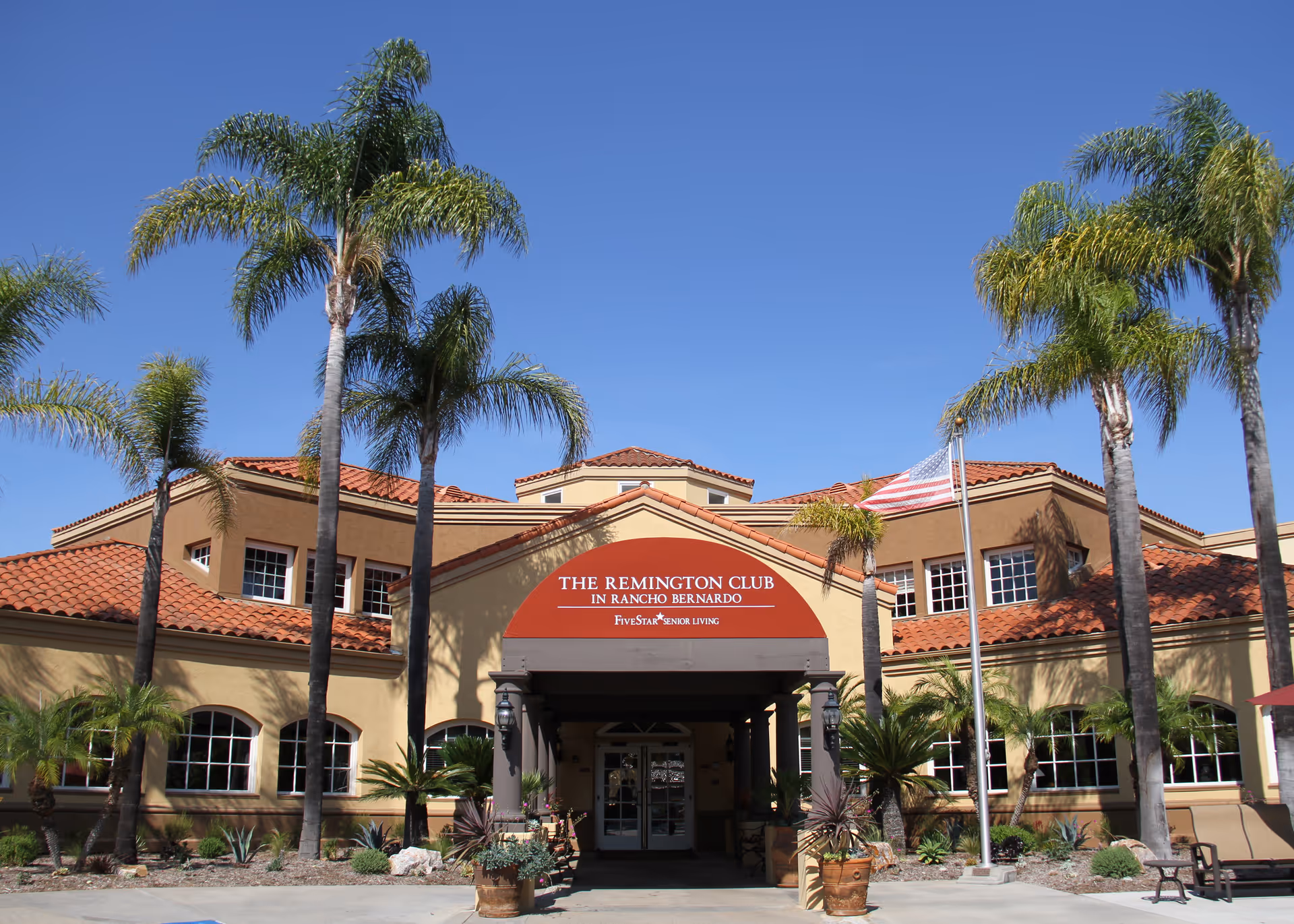 Front exterior view of The Remington Club senior living facility with palm trees, a clear blue sky, and an American flag on a flagpole near the entrance.