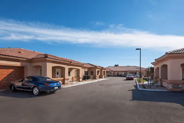 View of a senior living facility courtyard with single-story buildings featuring terracotta tiled roofs and beige walls. There are parked cars along the paved driveway under a clear blue sky.