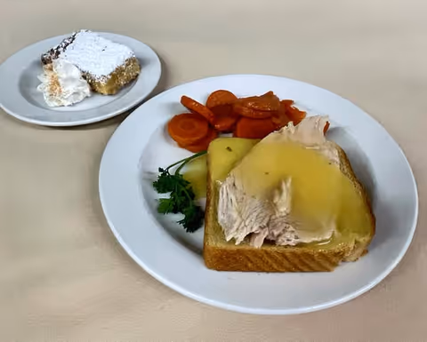 A meal served on a white plate consisting of sliced turkey with gravy on a slice of bread, cooked carrots, and a sprig of parsley. Next to it is a small dessert plate with a powdered sugar-topped square pastry and a dollop of whipped cream, all placed on a beige tablecloth.