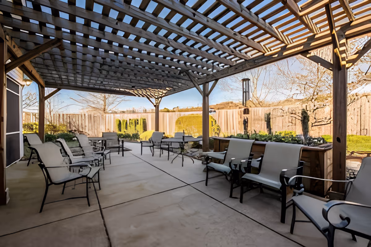 Outdoor covered patio area with multiple cushioned chairs arranged along the sides. The patio has a wooden pergola overhead providing partial shade. Surrounding the patio are landscaped garden areas with bushes and trees, and a wooden fence encloses the space.