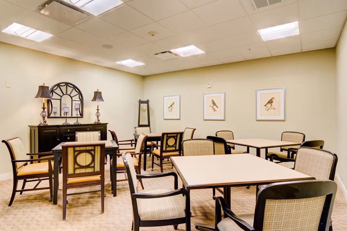 A well-lit room with several square tables surrounded by cushioned chairs. The walls are light beige and decorated with framed pictures of birds. There is a black sideboard with two lamps and a decorative mirror above it. The room has a beige carpet and a ceiling with fluorescent lights.