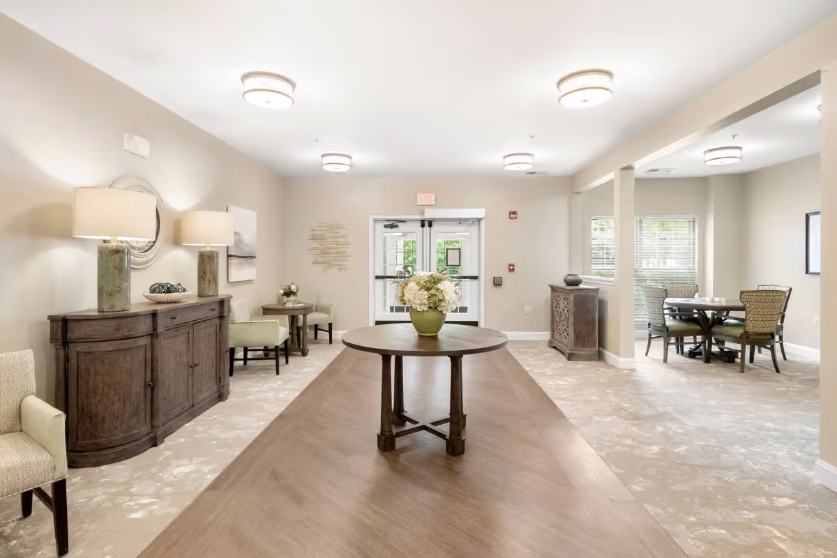 A bright and spacious common area in a senior living facility with beige walls and carpeted floors. The room features a long wooden table with a round table on top holding a green vase with white flowers. Along the left wall is a wooden sideboard with two lamps and decorative items. There are several seating areas with chairs and small tables, and large windows letting in natural light. The double glass doors at the far end lead outside.
