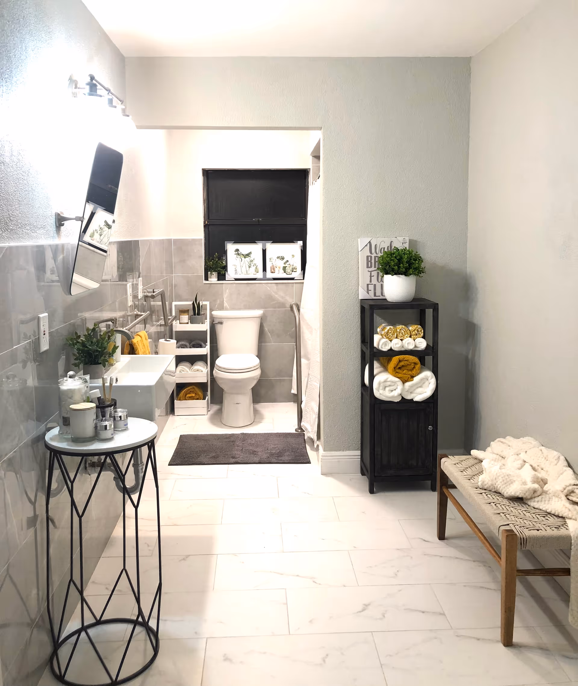 A modern bathroom with light gray tiled walls and white marble floor tiles. The room features a white toilet, a white sink with a mirror above it, and a small black shelving unit holding rolled towels and a potted plant. There is a small round table with candles and a plant on the left side, and a wooden bench with a knitted blanket on the right side. The bathroom is well-lit with a window above the toilet and a light fixture above the mirror.