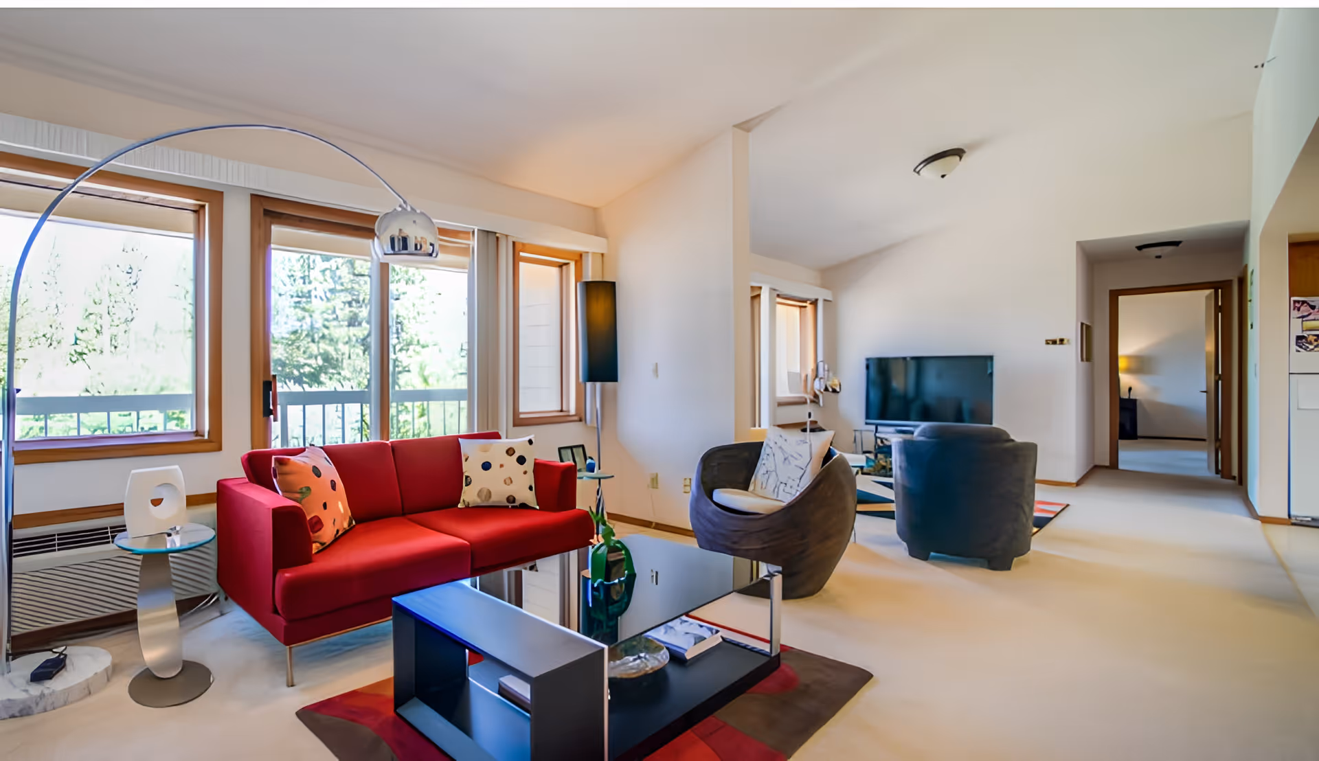 A bright living room with large windows showing greenery outside. The room features a red sofa with patterned pillows, a modern arc floor lamp, a glass coffee table with decorative items, a round chair with a cushion, and a TV on a stand. The walls are white, and the carpet is light-colored. There is an open doorway leading to another room.
