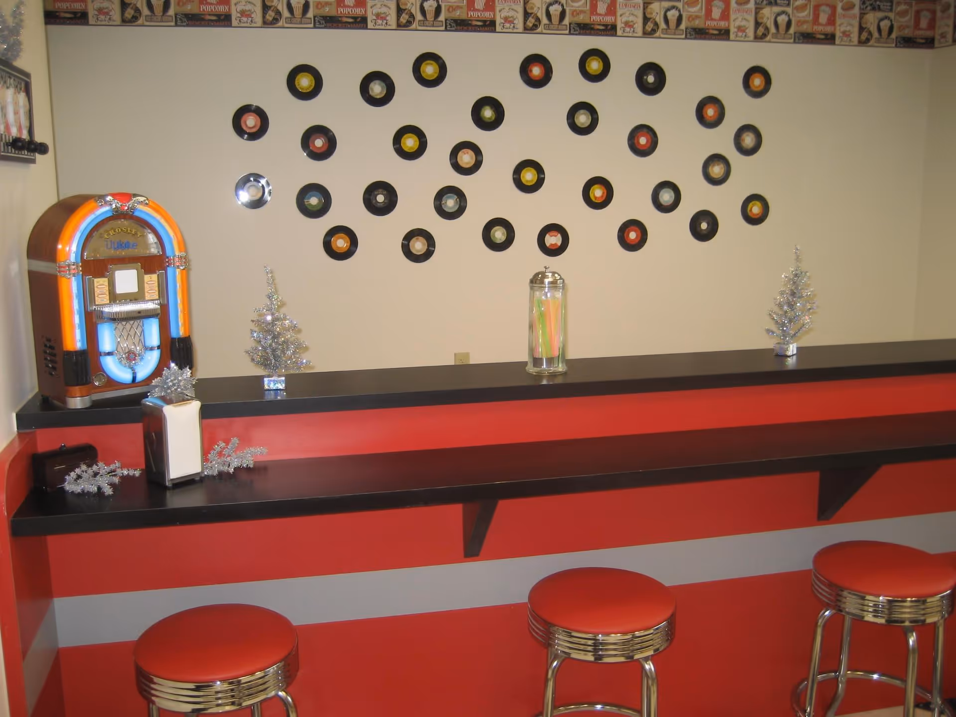 A retro-style diner counter with three red cushioned stools. The counter is black with a red front panel. On the counter, there is a small jukebox, two small silver decorative trees, a napkin holder, and a glass jar with colorful straws. The wall behind the counter is decorated with small vinyl records arranged in a pattern and a border of vintage popcorn-themed wallpaper at the top.