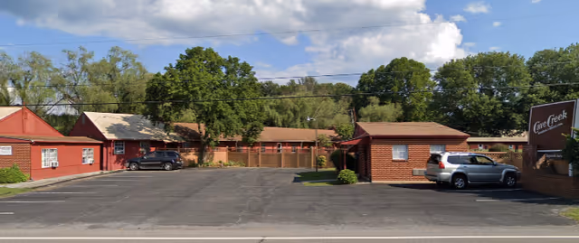 Exterior view of Cave Creek Assisted Living facility showing a parking lot with two parked cars, several single-story buildings with red and brown exteriors, trees in the background, and a sign on the right side that reads 'Cave Creek Assisted Living'.