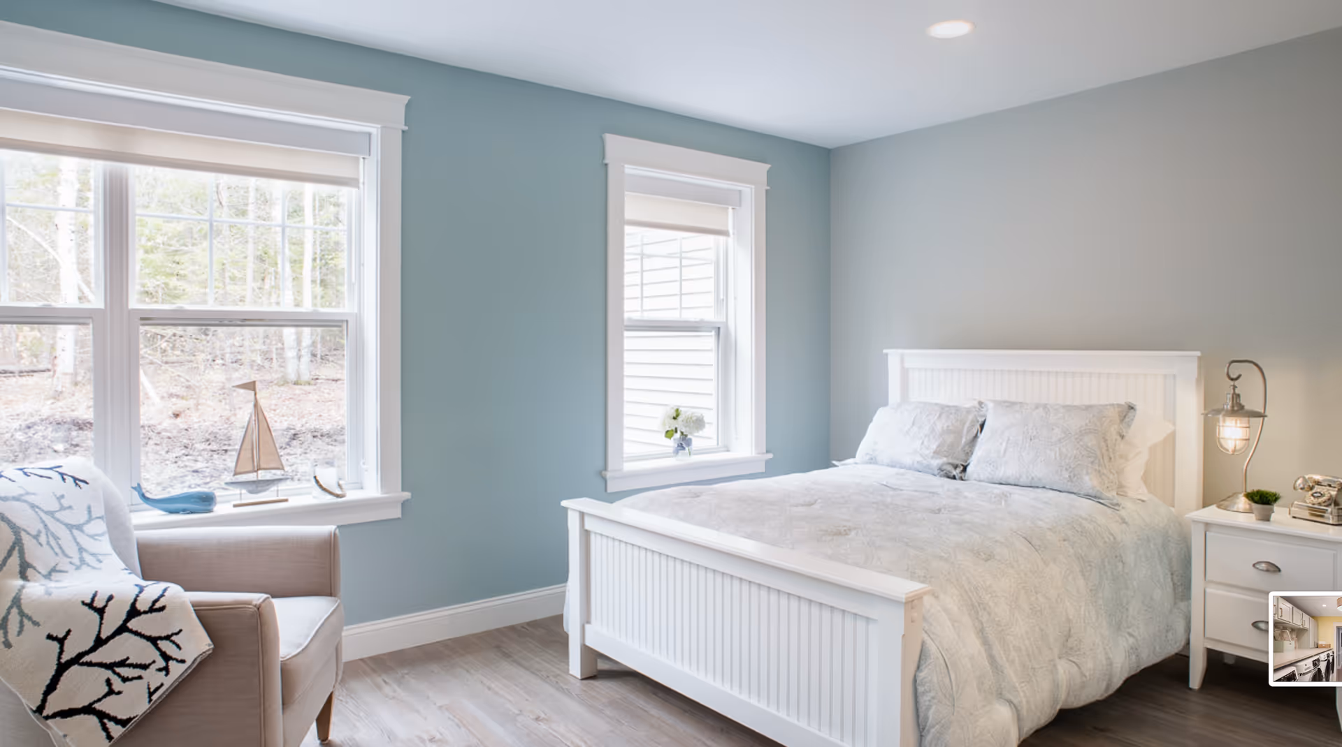 Sunlit bedroom with a white wooden bed, bedside table and lamp, armchair by large windows, and soft blue-gray walls.